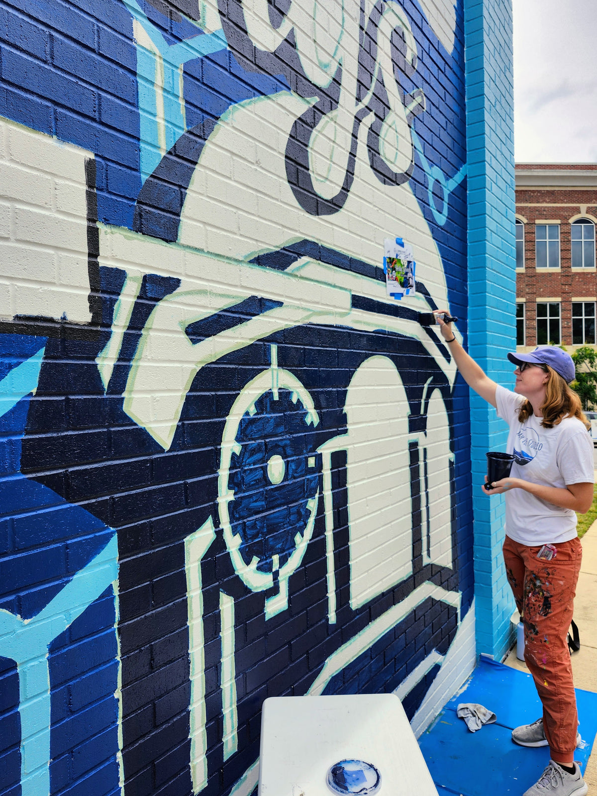 Person painting a large mural of a helmet on a wall with blue, white, and black colors.