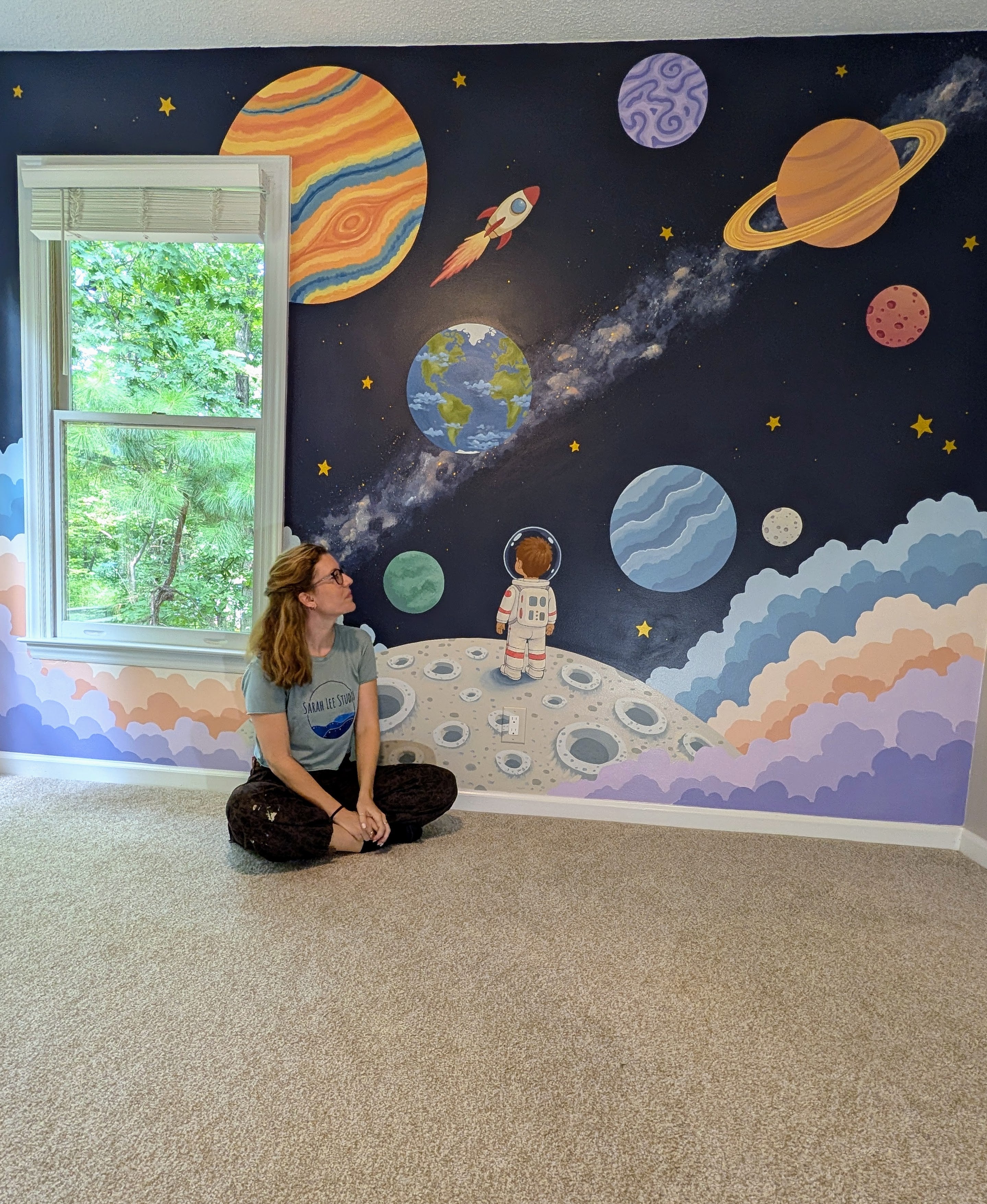 A child's room featuring a large, colorful space-themed mural on the wall with planets, stars, a rocket, and an astronaut. A young girl with long light brown hair sits on the beige carpet on the floor, looking up at the mural.