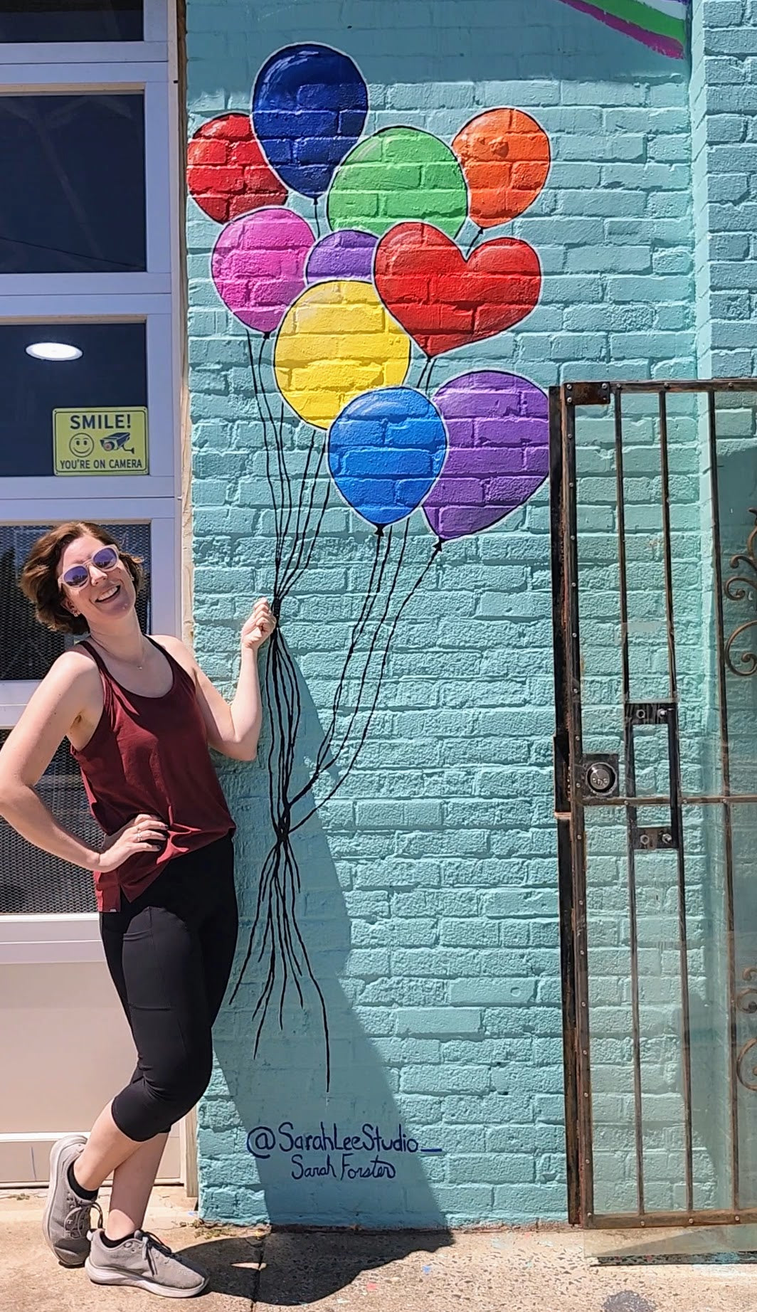 A woman in a maroon tank top and black leggings poses next to a light blue brick wall featuring a colorful mural of heart and balloon shapes.